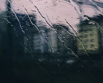Submerged Rain-streaked train window with blurred city buildings barely visible behind dark green and gray tones.