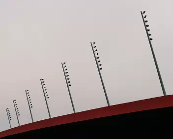 Measured Ascent Minimalist architectural photograph featuring a line of stadium floodlights ascending above a red roof against a soft grey sky - a study in balance, rhythm, and...