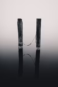 Tethered Quiet Two weathered wooden posts stand in calm water with a rope loosely connecting them, reflected in the still surface.