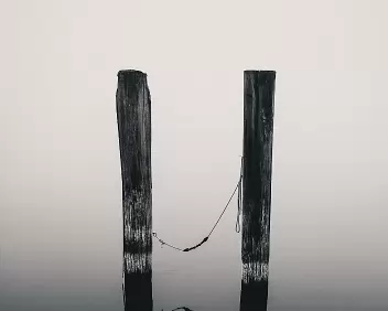 Tethered Quiet Two weathered wooden posts stand in calm water with a rope loosely connecting them, reflected in the still surface.