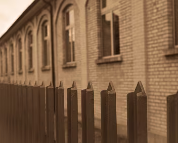 Fence Line A metal fence runs parallel to a brick facade, its repetition echoing the rhythm of the windows behind it. The shallow focus compresses distance, turning...