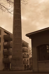 Industrial Spine An old brick chimney stands between former industrial buildings and newly built apartments. Once central to production, it now remains as a vertical witness —...